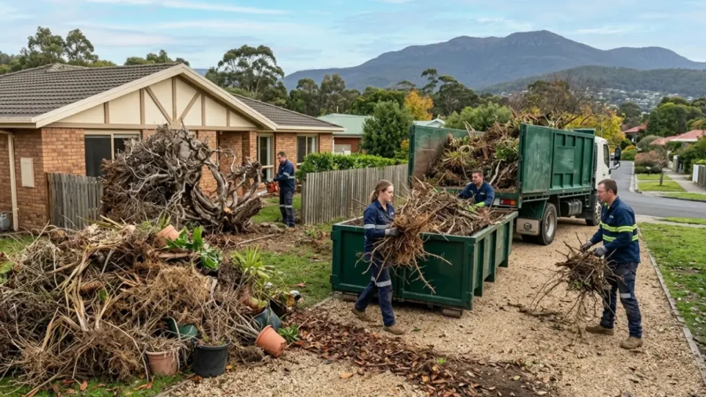 Workers clearing large fallen tree branches and debris into a dumpster outside a house after a storm to make the garden clear from waste.
